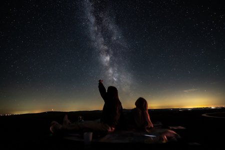 2 Mädchen sitzen am Boden und schauen in den Sternenhimmel, eine zeigt in den Sternenhimmel