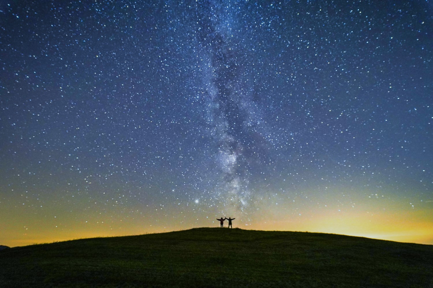 2 Personen fassen sich an der Hand, stehen auf einem Berg und schauen in den Sternenhimmel.