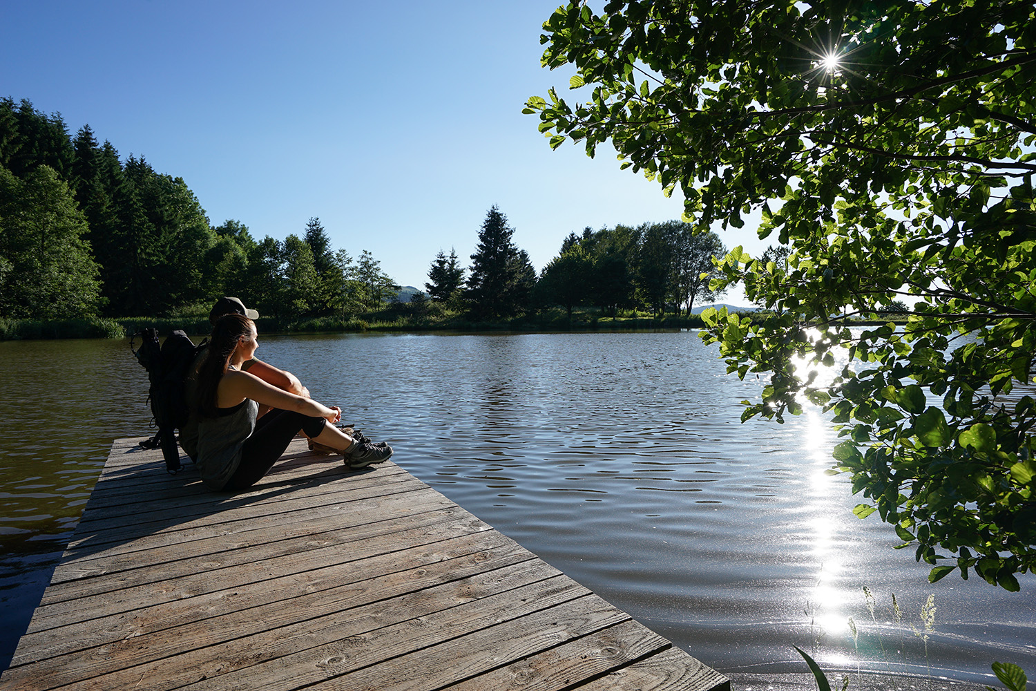 2 Wanderer sitzen auf dem Holzsteg am Naturweiher Reulbach und genießen die Sonne